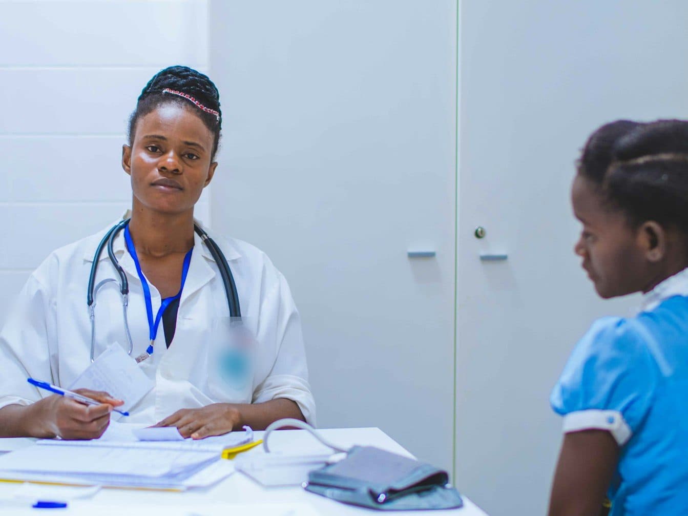 A clinician writing up notes at a consulting-room desk, with a child patient seated opposite. Paediatric and neonatal intensive care, for children and newborn babies, are planned services at TEMCO, opening 2028.