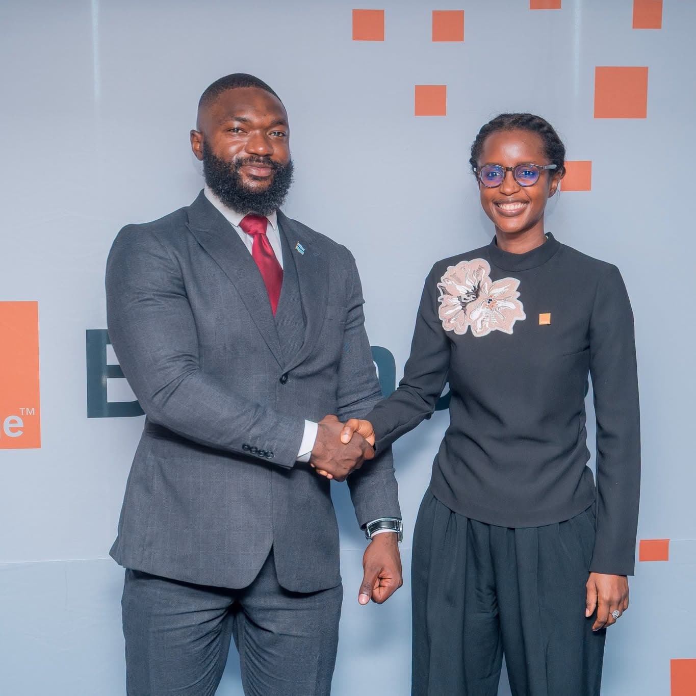 Two people shaking hands in front of a branded backdrop, facing the camera.