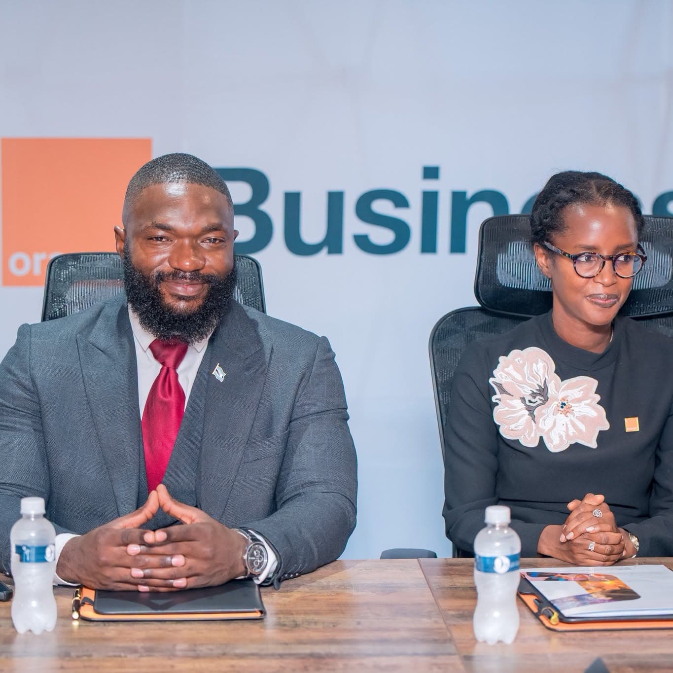 A man and a woman seated side by side at a boardroom table, facing the camera.